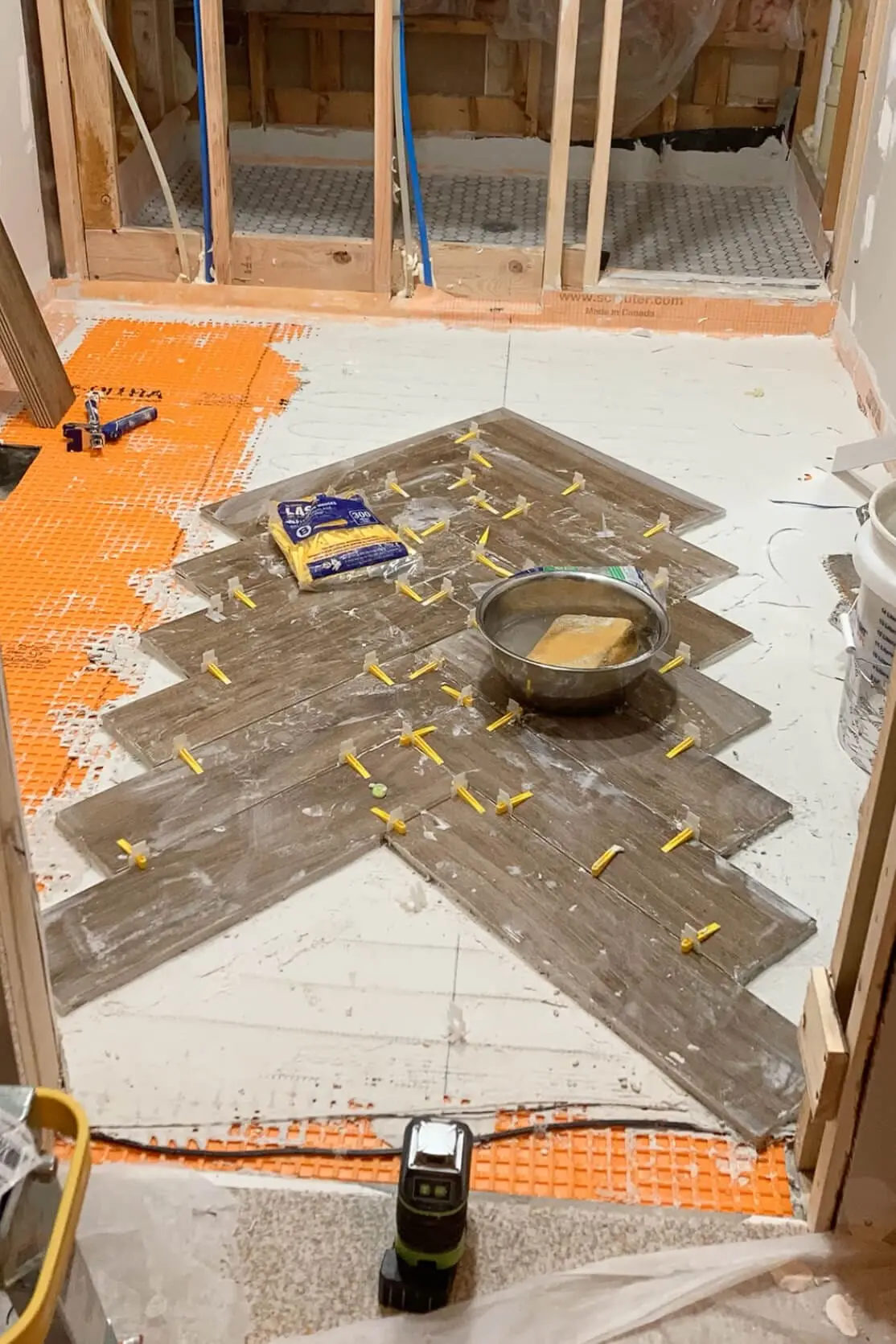 A herringbone tile pattern partially completed on a bathroom floor with yellow spacers, tools, and a sponge in a metal bowl nearby for easy cleanup during installation.
